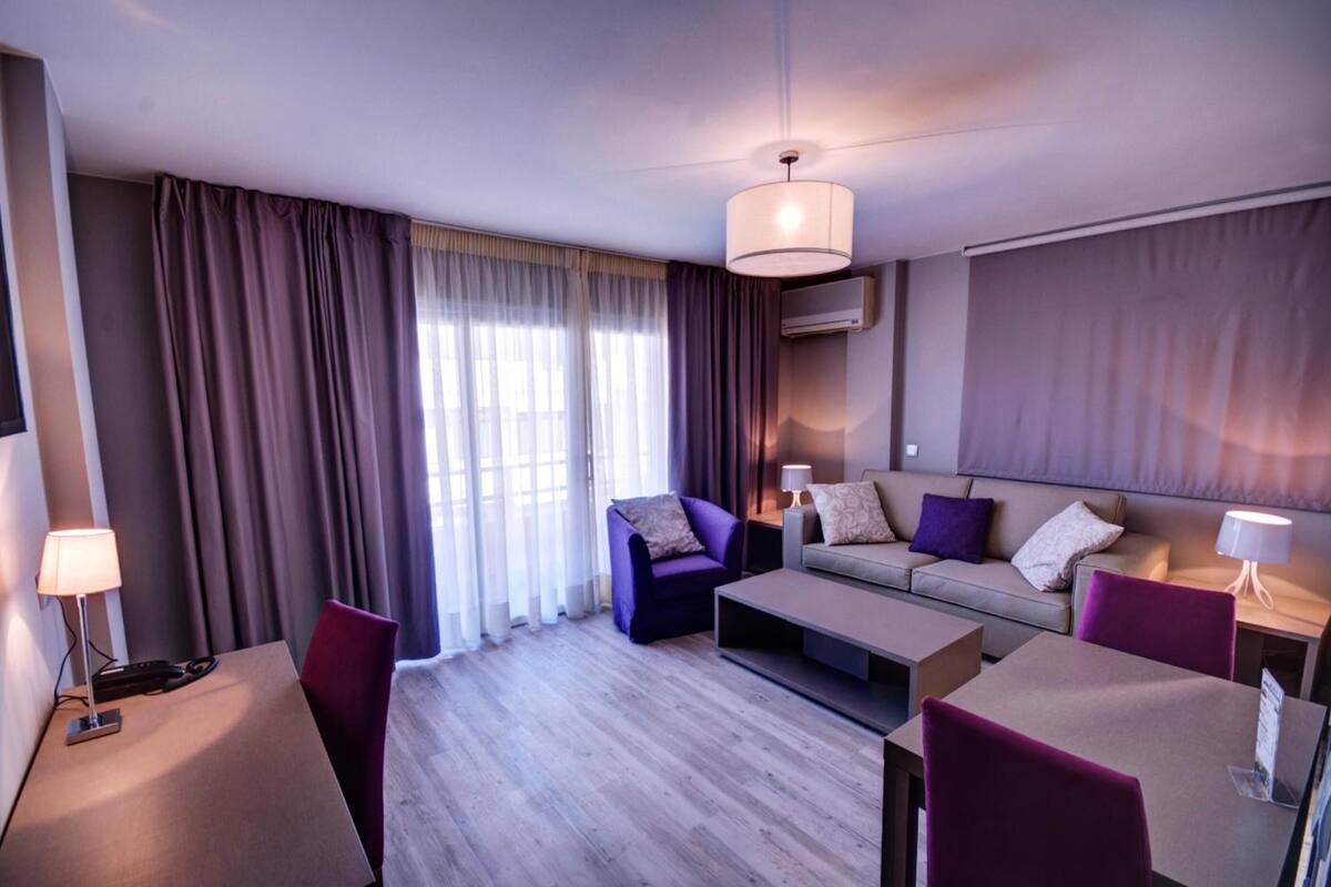 A modern hotel room with a sofa, armchair, and a dining table.