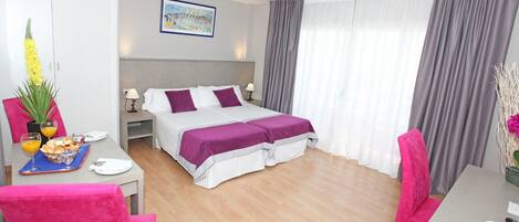 A hotel room with a bed, a desk with a chair, a dining table with chairs, and a large window with curtains.