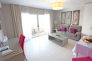 A modern living room with a sofa, coffee table, and dining area. There is a balcony with curtains and a table with fruit and a drink.