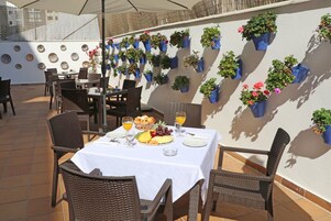 Een terras buiten met rieten meubels, een tafel gedekt met een fruitschaal en drankjes, en een muur versierd met hangende plantenbakken.