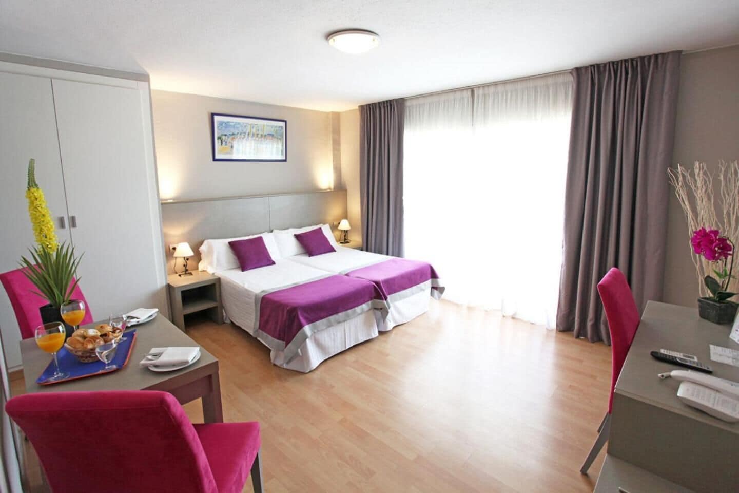 A hotel room with a bed, bedside tables, a desk, a chair, a dining table, and a potted plant.
