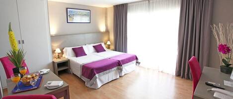A hotel room with a bed, bedside tables, a desk, a chair, a dining table, and a potted plant.