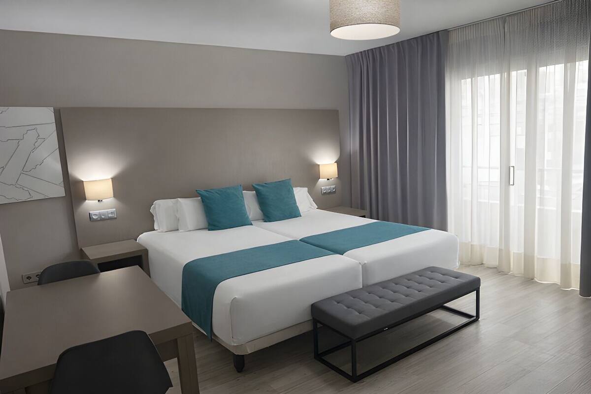A modern hotel room with a large bed, bedside tables, a desk, and a bench.