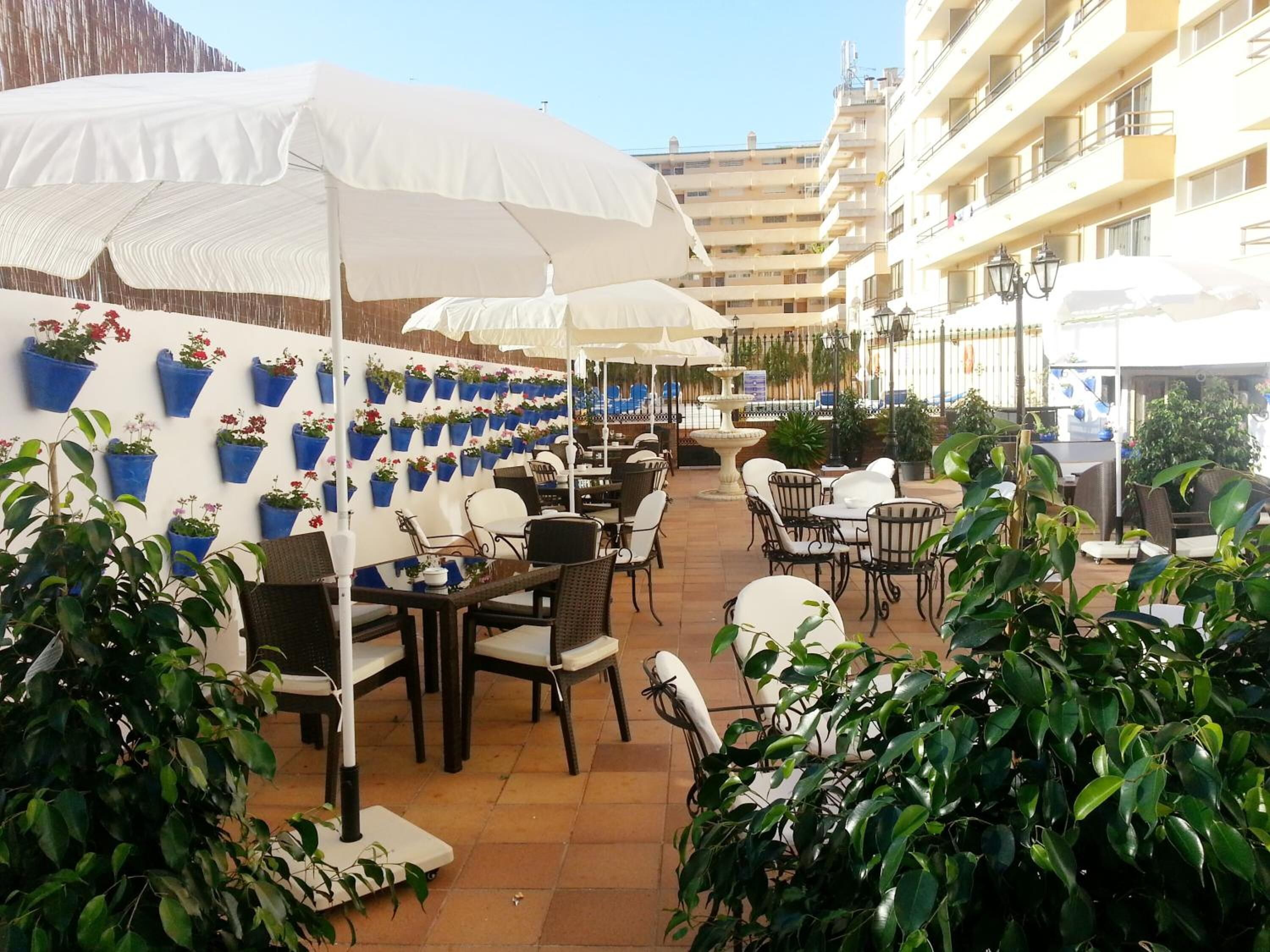 Een buitenterras met tafels en stoelen, witte parasols en plantenbakken aan de muren.