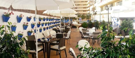 Een buitenterras met tafels en stoelen, witte parasols en plantenbakken aan de muren.