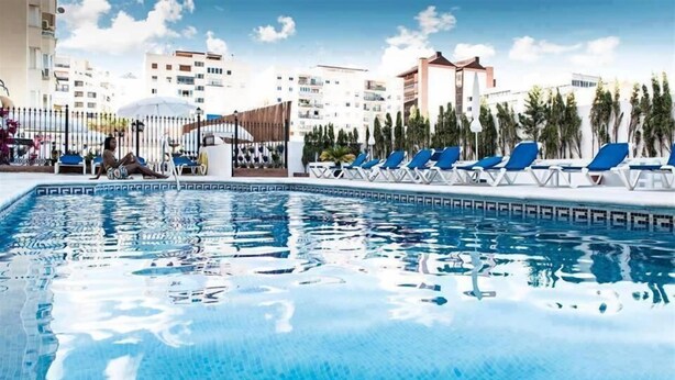 A swimming pool with lounge chairs and umbrellas, surrounded by buildings and palm trees. - Hotel El Faro Marbella (Marbella)