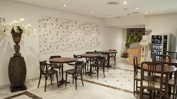 Een hotellobby met meerdere houten tafels en stoelen, een grote decoratieve vaas met bloemen, een behang met patroon en een automaat.