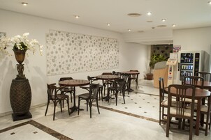 Een hotellobby met meerdere houten tafels en stoelen, een grote decoratieve vaas met bloemen, een behang met patroon en een automaat.