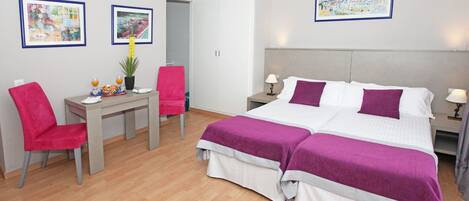 A hotel room with a double bed, a desk with a chair, a small table with a plant, and two framed pictures on the walls.
