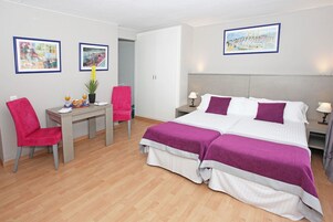 A hotel room with a double bed, a desk with a chair, a small table with a plant, and two framed pictures on the walls.