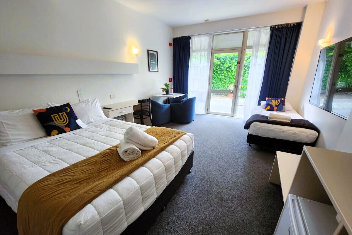 Habitación de hotel con una cama grande, una cama más pequeña, un escritorio y una silla. Se ve una ventana con cortinas y vistas al verde exterior.