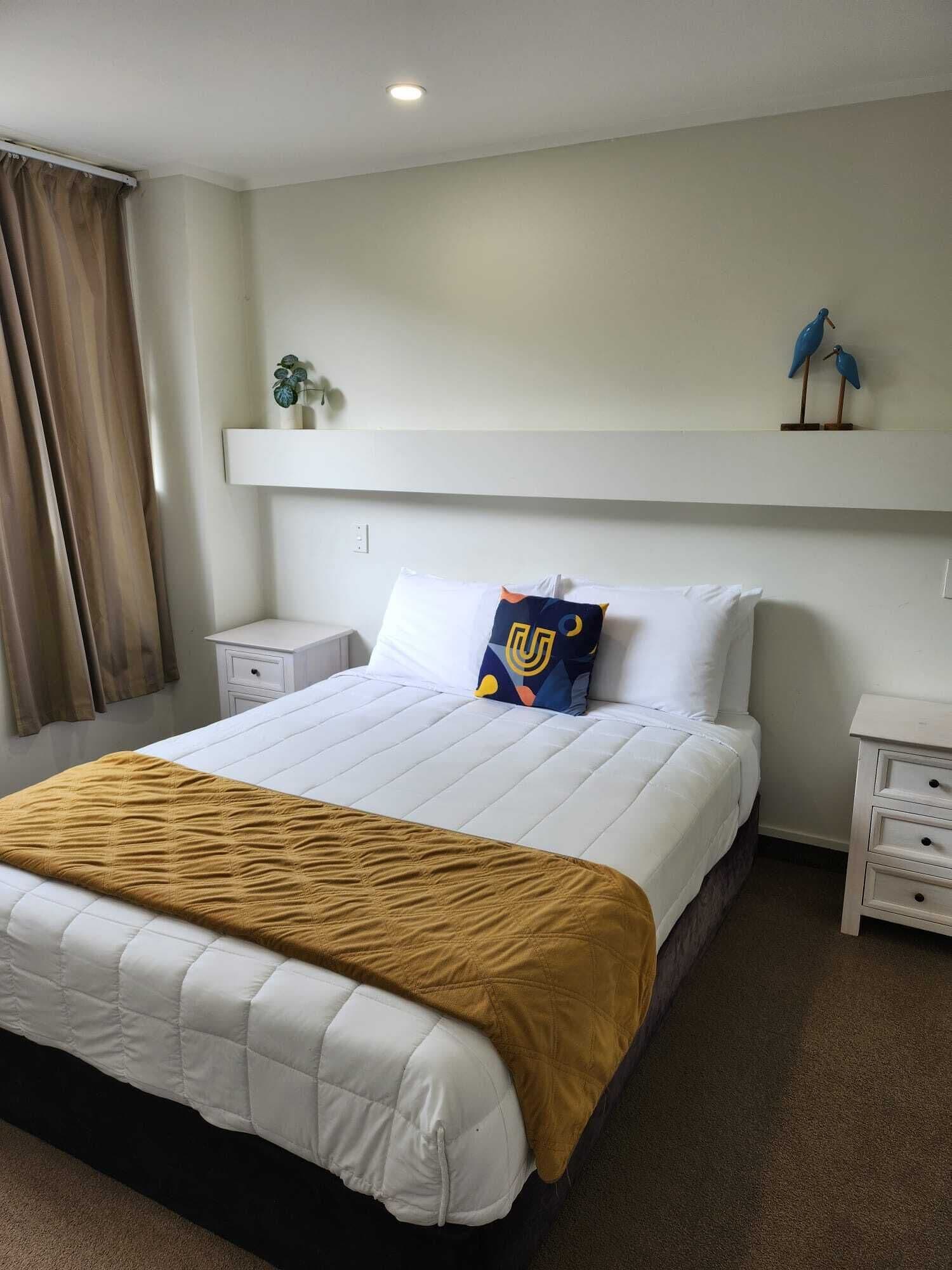 A neatly arranged hotel room with a large bed, a bedside table, a nightstand, a shelf with decorative items, and a window with curtains.