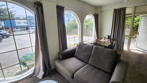 A living room with a sofa, a table, and a ceiling fan. Large windows with curtains are visible. -  U Studios Paraparaumu, SureStay Collection by BW (Paraparaumu)