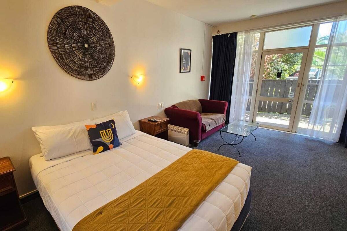 Una habitación de hotel con cama, una silla, una lámpara de pared y un ventanal con cortinas.