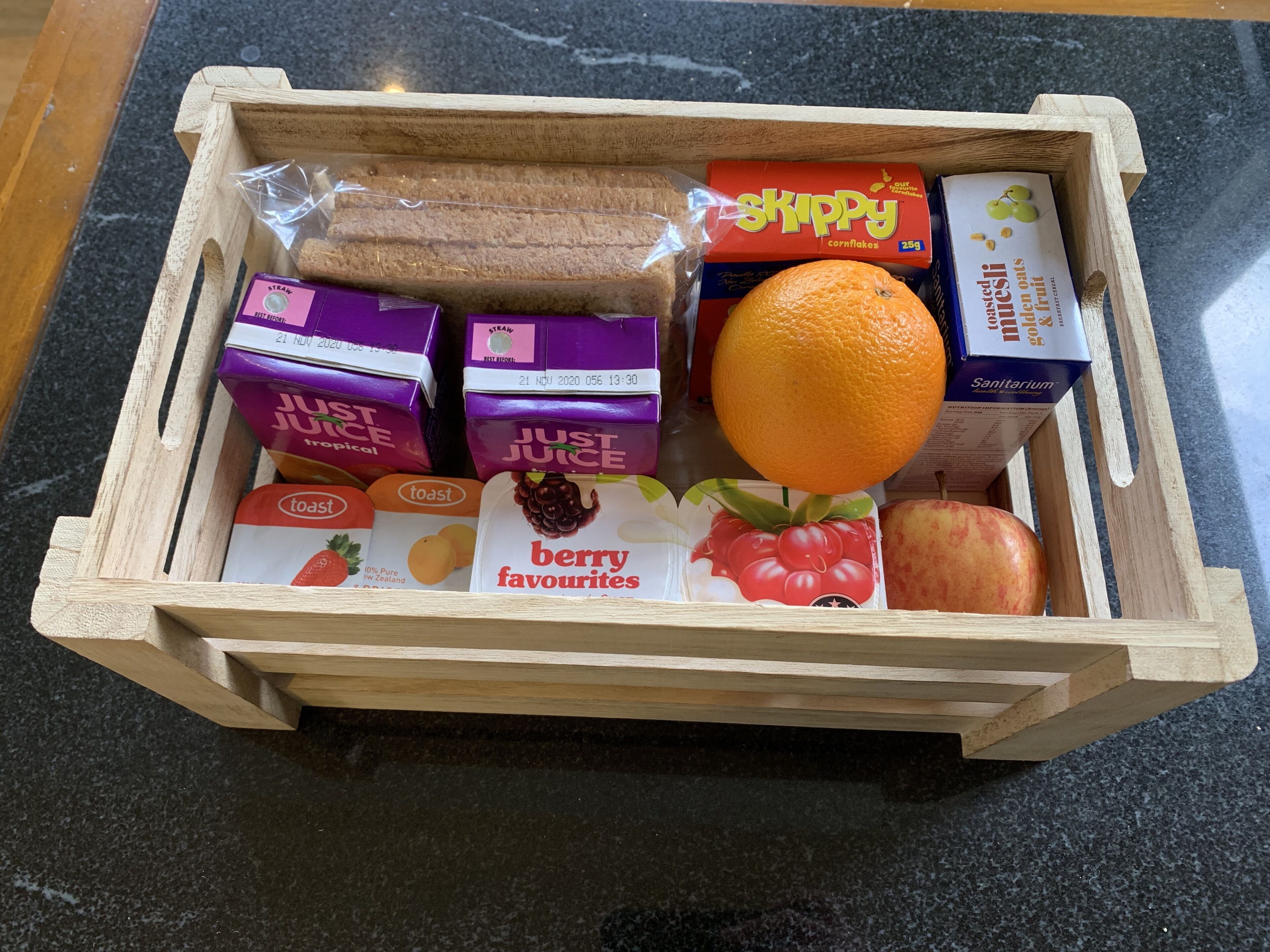 Una cassa di legno contenente diversi alimenti, tra cui cereali in scatola, succo di frutta, pane tostato e un'arancia intera.