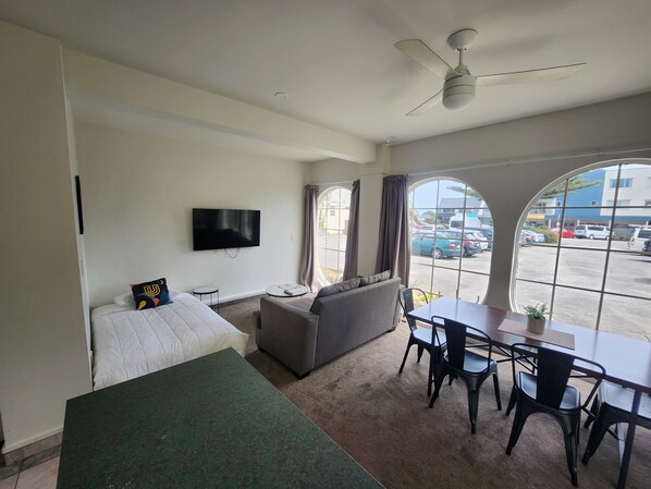 A hotel room with a bed, a sofa, a TV, a dining table with chairs, and a large window with a view of the parking lot. - U Studios Paraparaumu, SureStay Collection by BW (Paraparaumu)