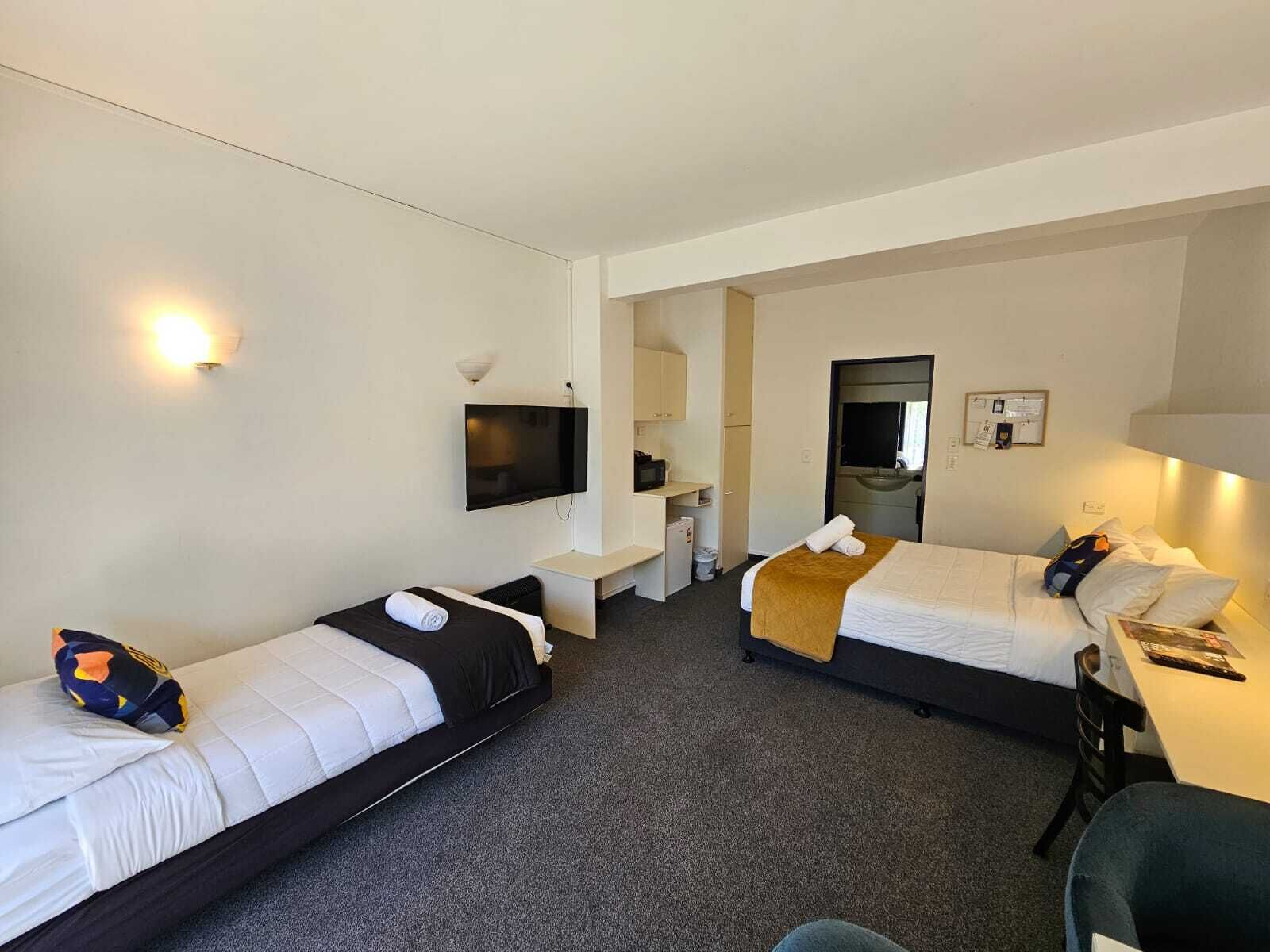 A hotel room with two single beds, a desk with a chair, a TV, a mirror, and a small kitchenette.