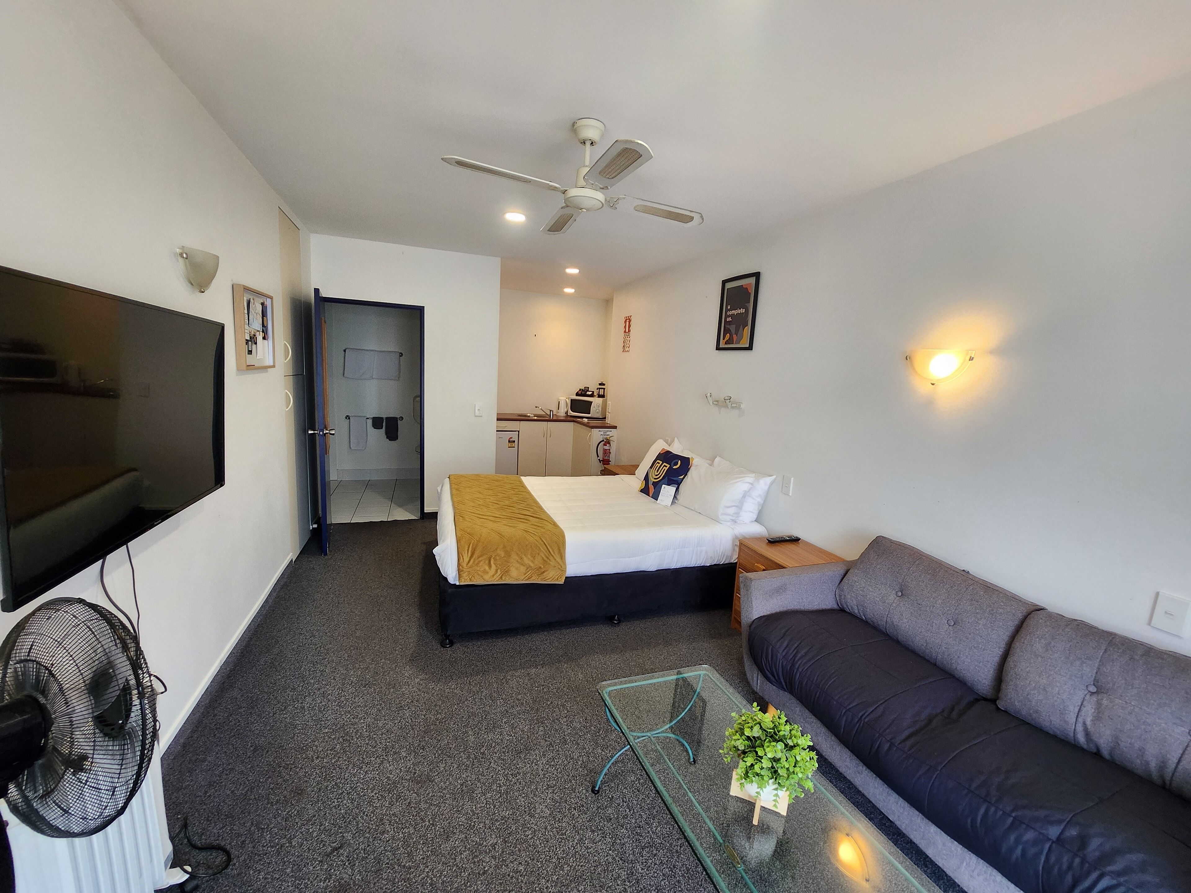 A hotel room with a bed, a sofa, a TV, a ceiling fan, a small kitchenette, and a bathroom.