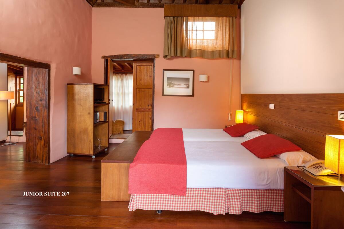 A junior suite with a wooden headboard, a bed with red bedding, a nightstand with a lamp, and a wooden wardrobe.
