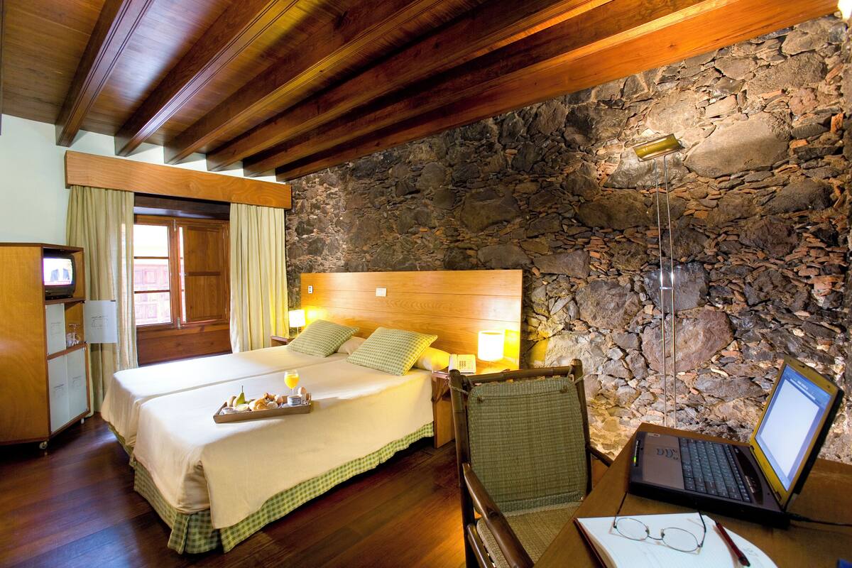 A hotel room with a stone wall, wooden ceiling beams, a bed with a tray of fruit, a laptop on a desk, and a chair.