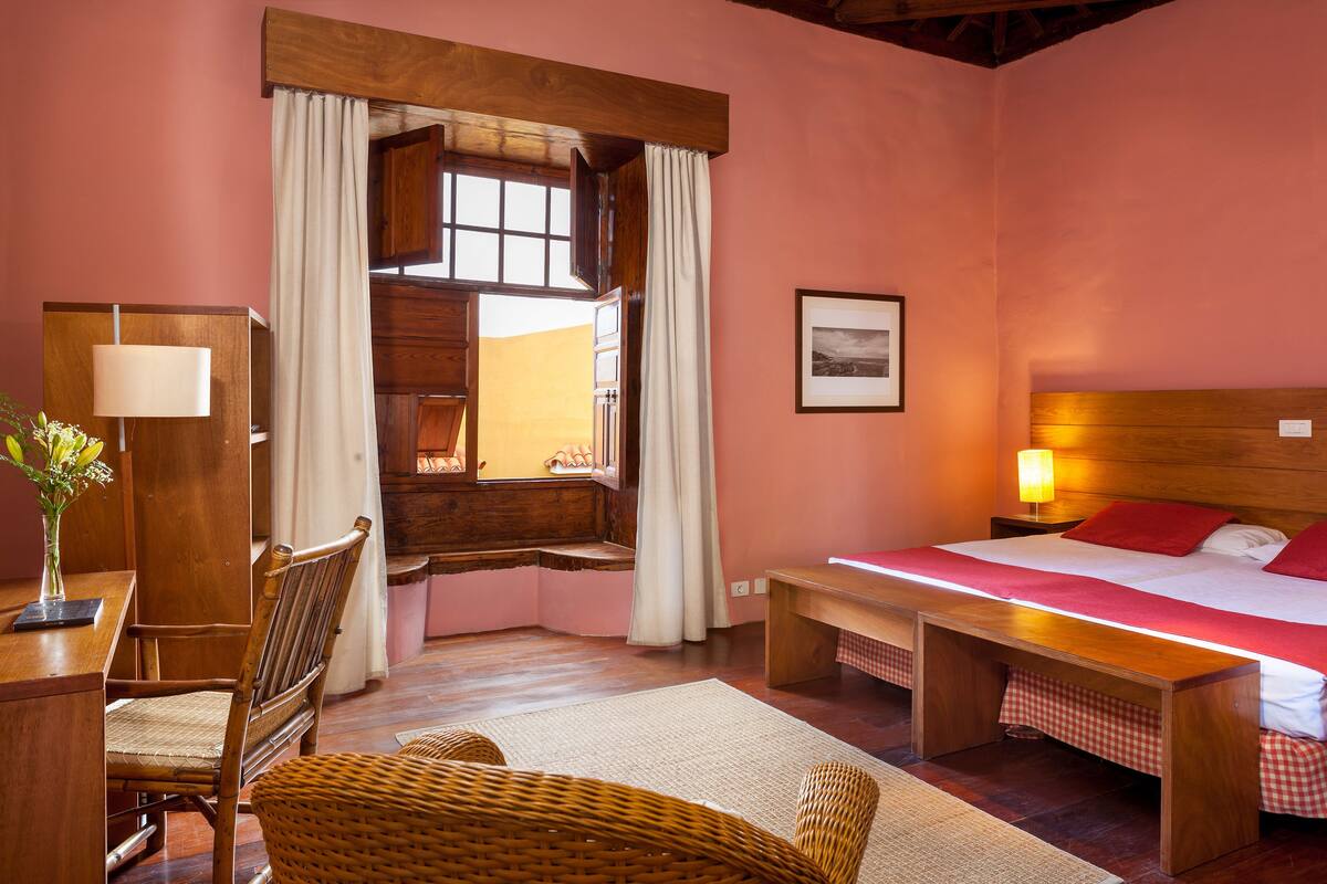 A cozy bedroom with a wooden bed, a desk with a lamp, a wicker chair, and a window with curtains.