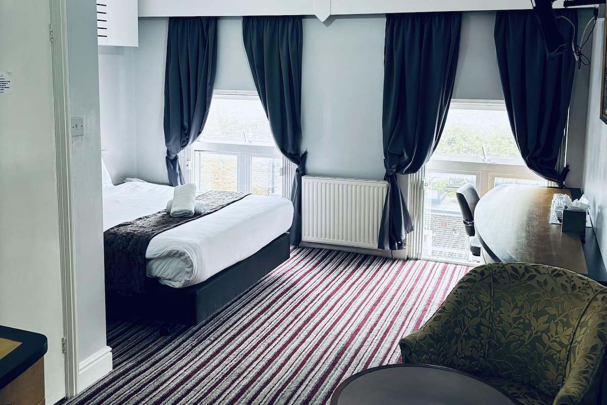Una camera d'albergo con un letto, una sedia, un tavolo, una ventola a soffitto e una finestra con tende.