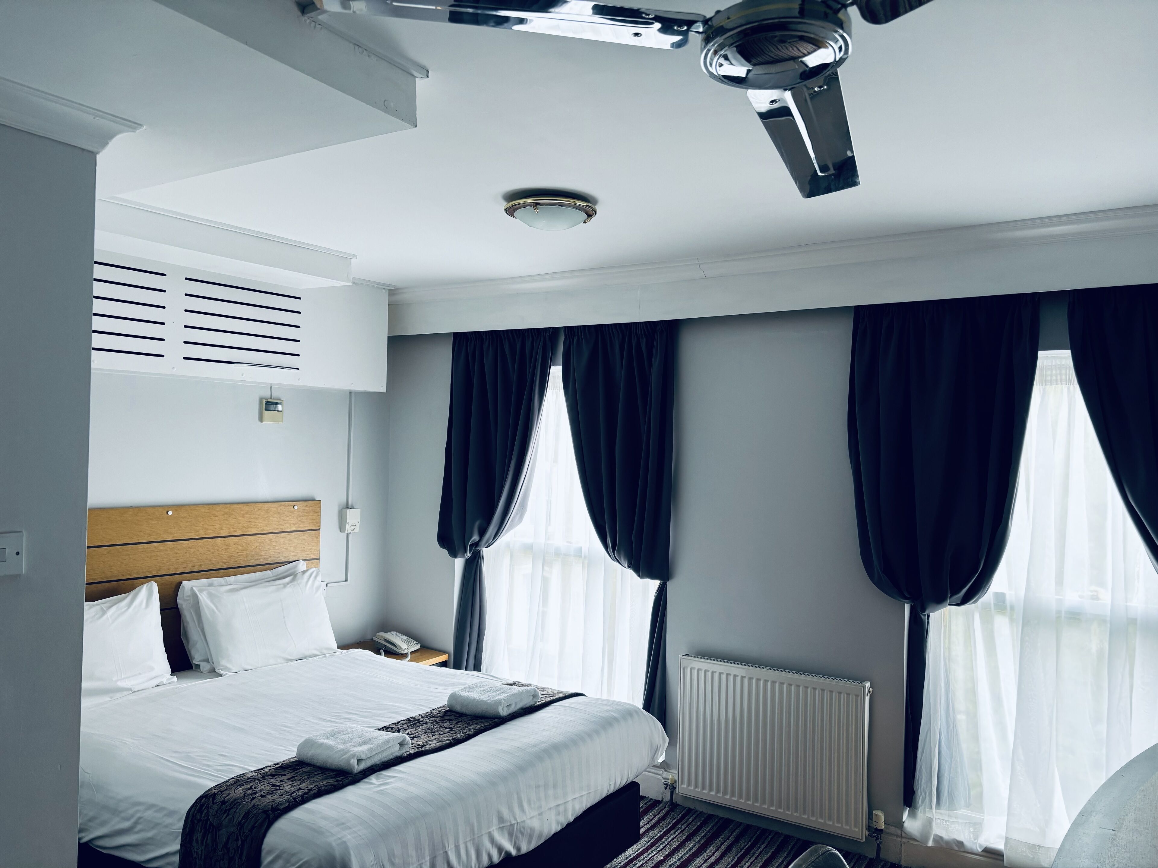 Habitación de hotel con cama, mesita de noche, radiador y ventilador de techo.
