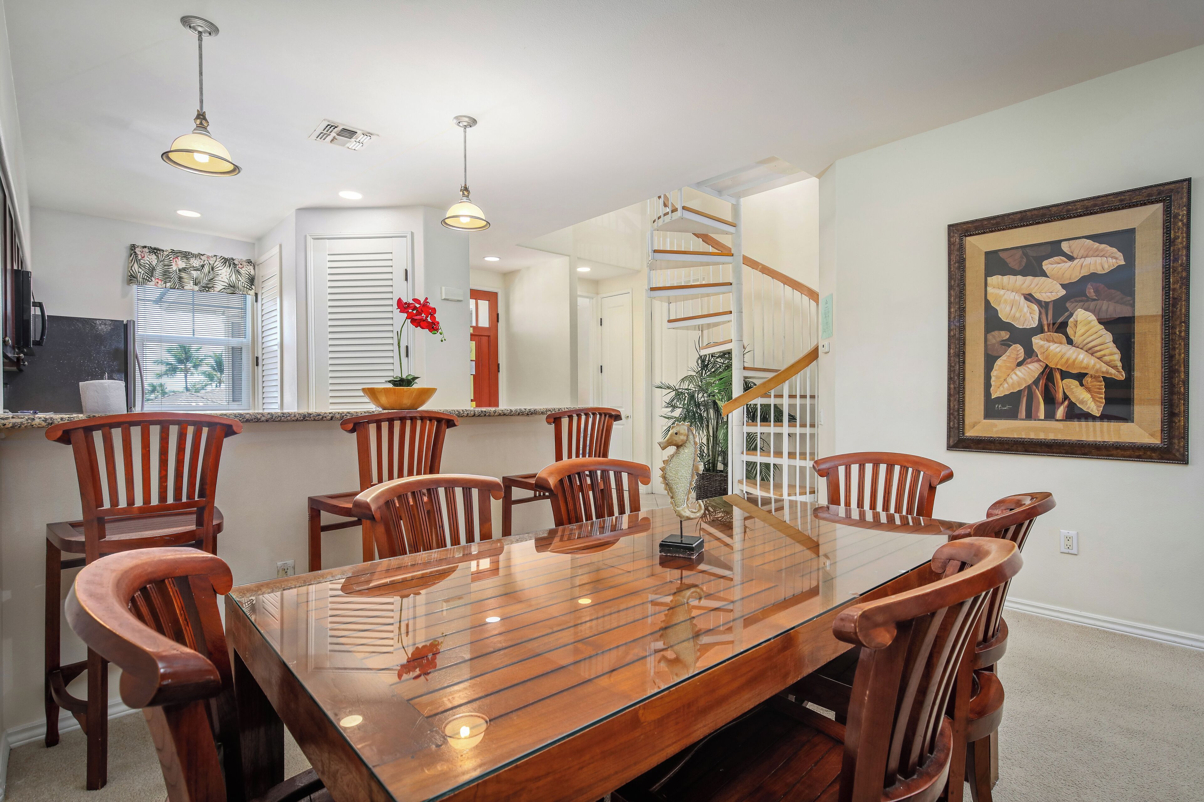 Un coin repas avec une table à plateau de verre, des chaises en bois, des luminaires suspendus, un escalier et un tableau encadré au mur.