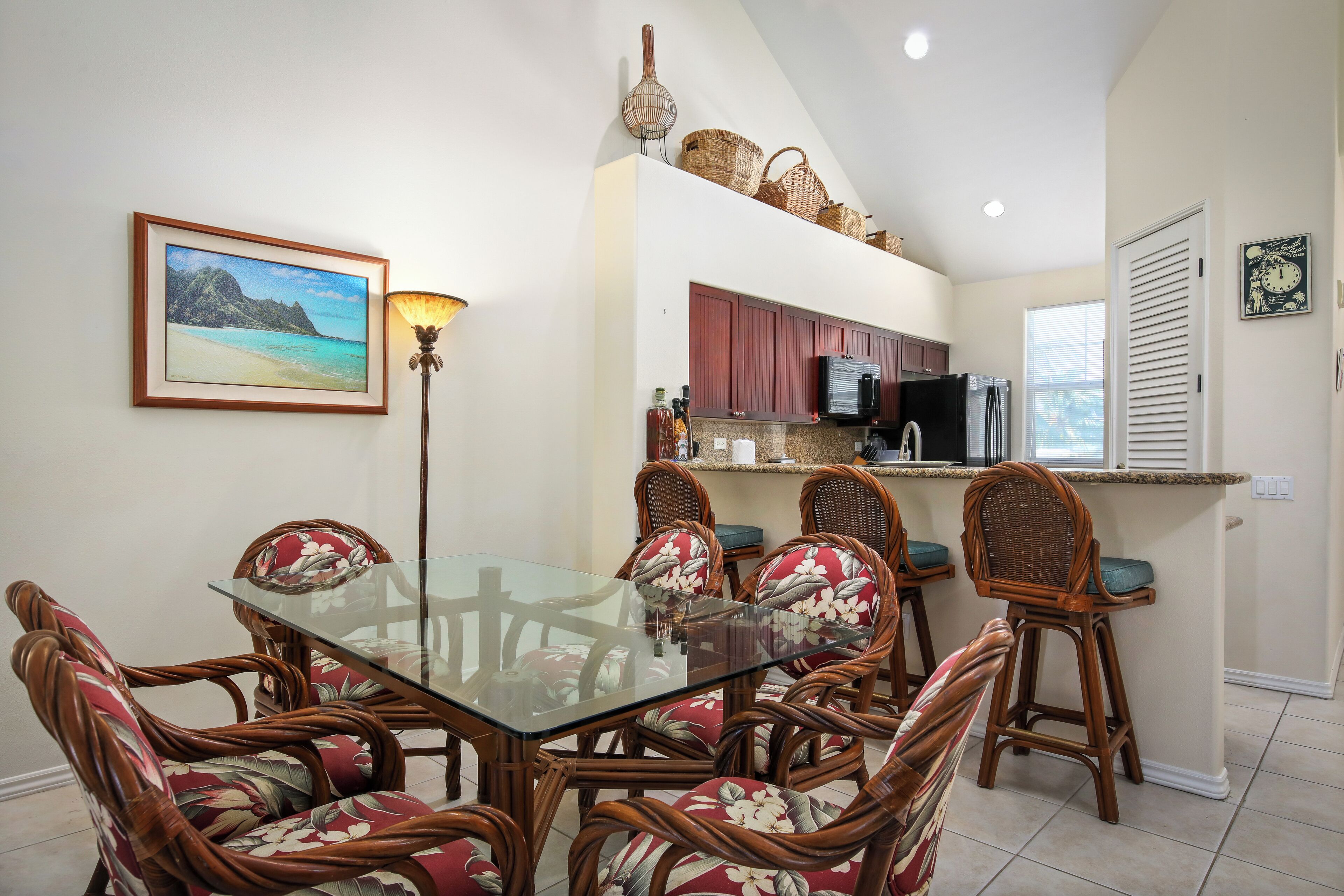 A dining area with a glass-top table surrounded by wicker chairs with floral upholstery, a framed picture on the wall, and a kitchen area with wooden cabinets and a built-in refrigerator.