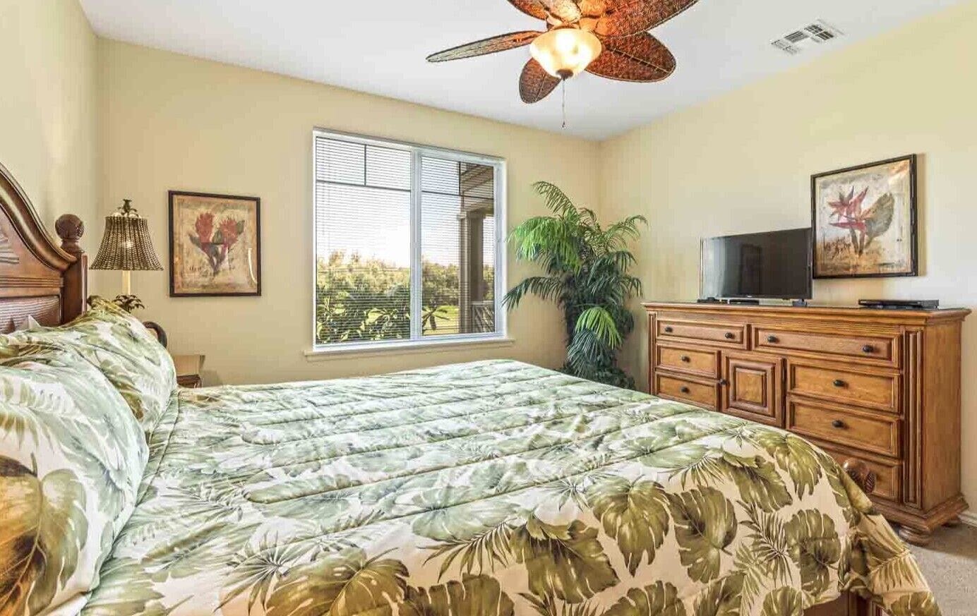 Un dormitorio con una cama, un aparador de madera, un televisor, un ventilador de techo y una ventana que ofrece vistas al verdor.