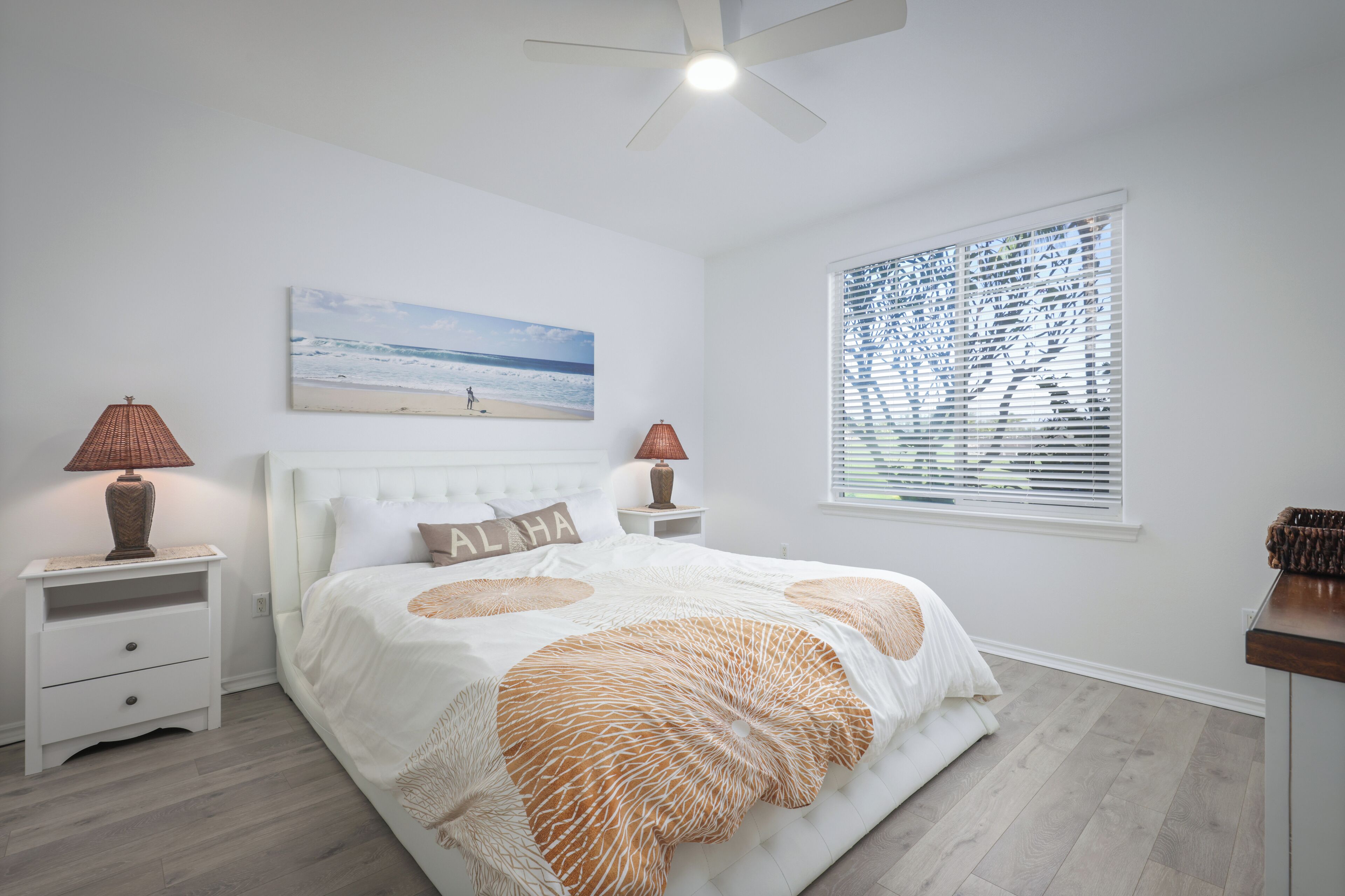 A bedroom with a bed, two nightstands, a ceiling fan, a beach-themed painting, and a window with blinds.