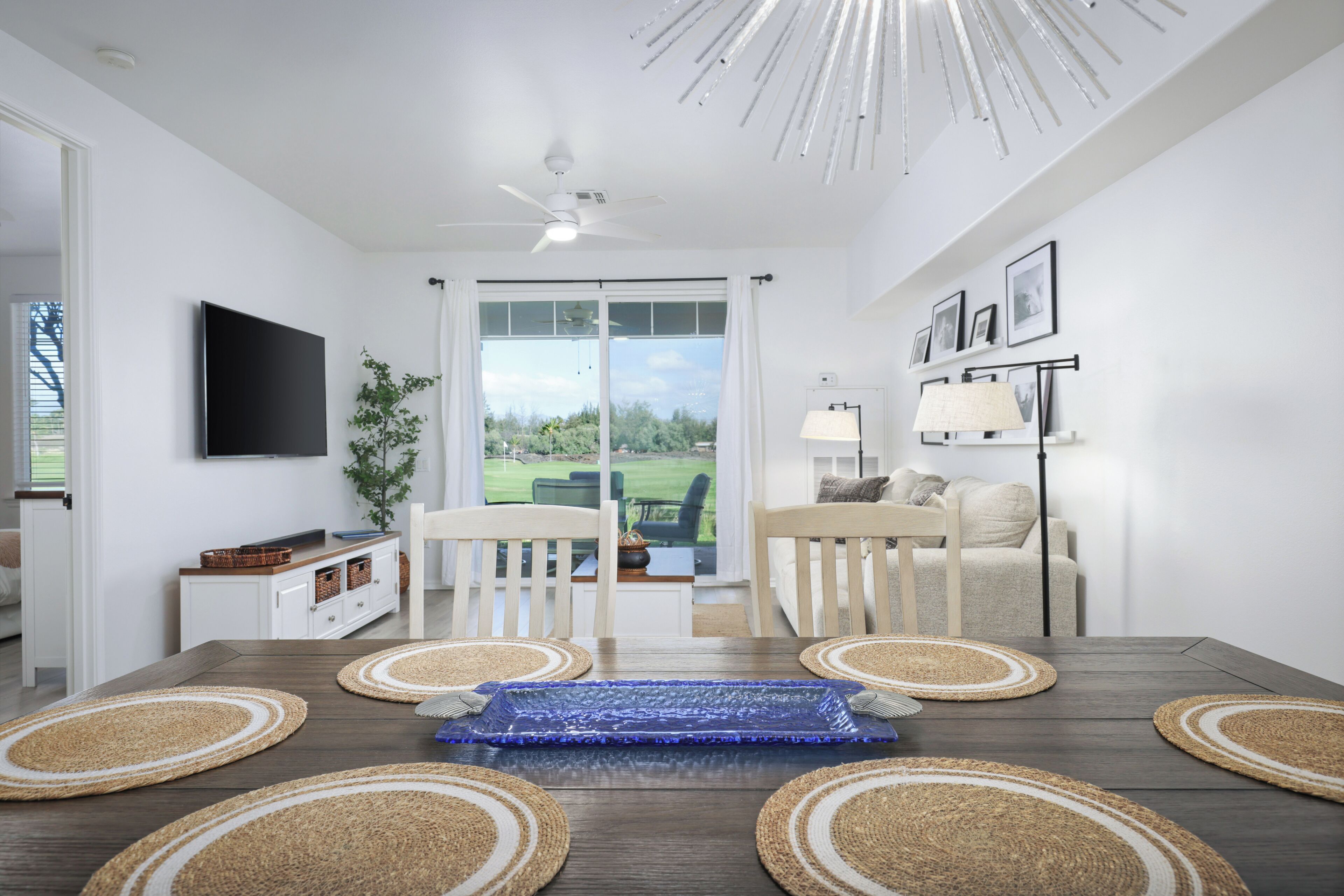 Un salón moderno con una mesa de comedor de madera oscura, sillas blancas, un televisor de pantalla plana y vistas a un paisaje verde a través de amplios ventanales.