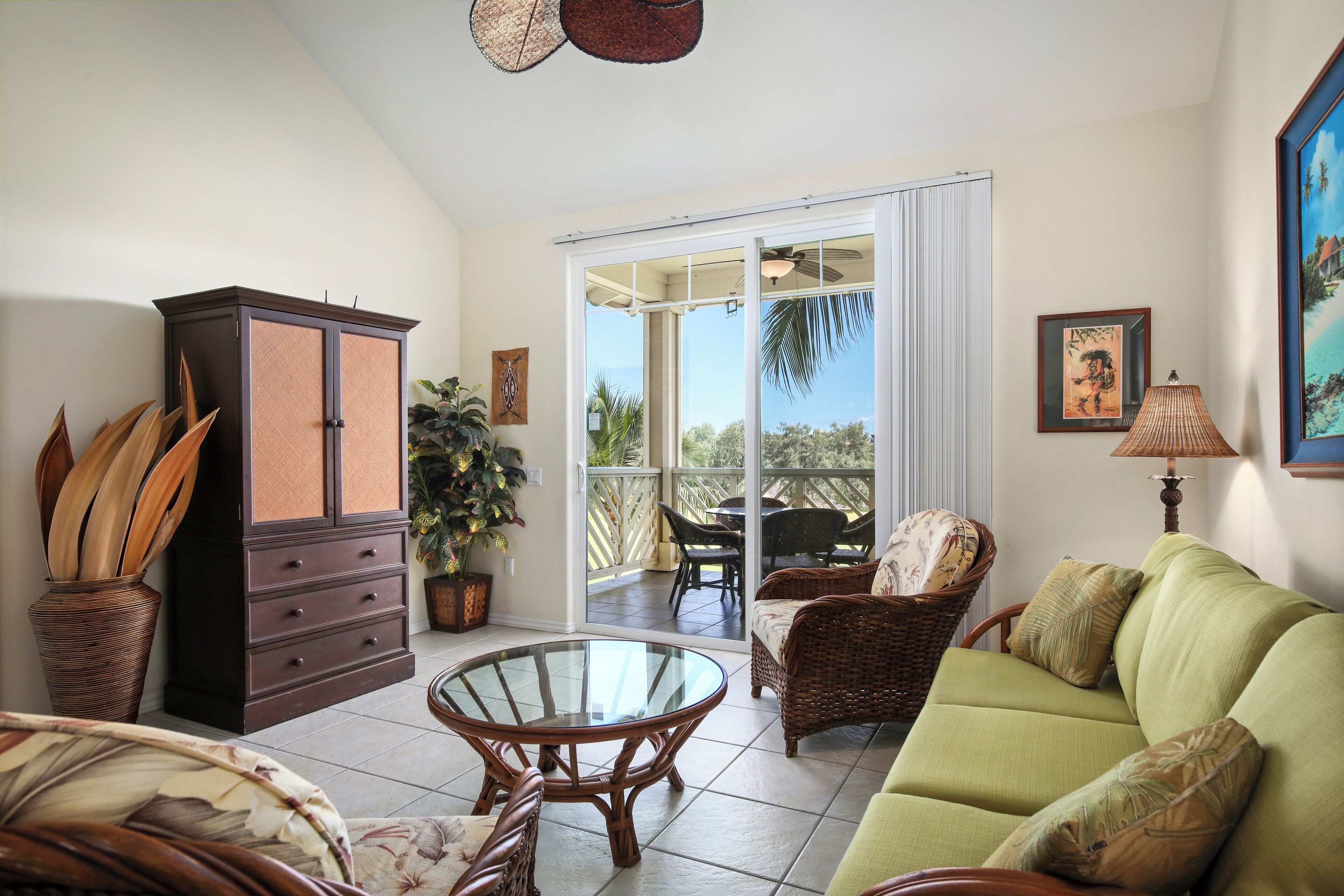 A living room with a green sofa, wicker chairs, a glass-top coffee table, a dark wooden cabinet, and a potted plant.