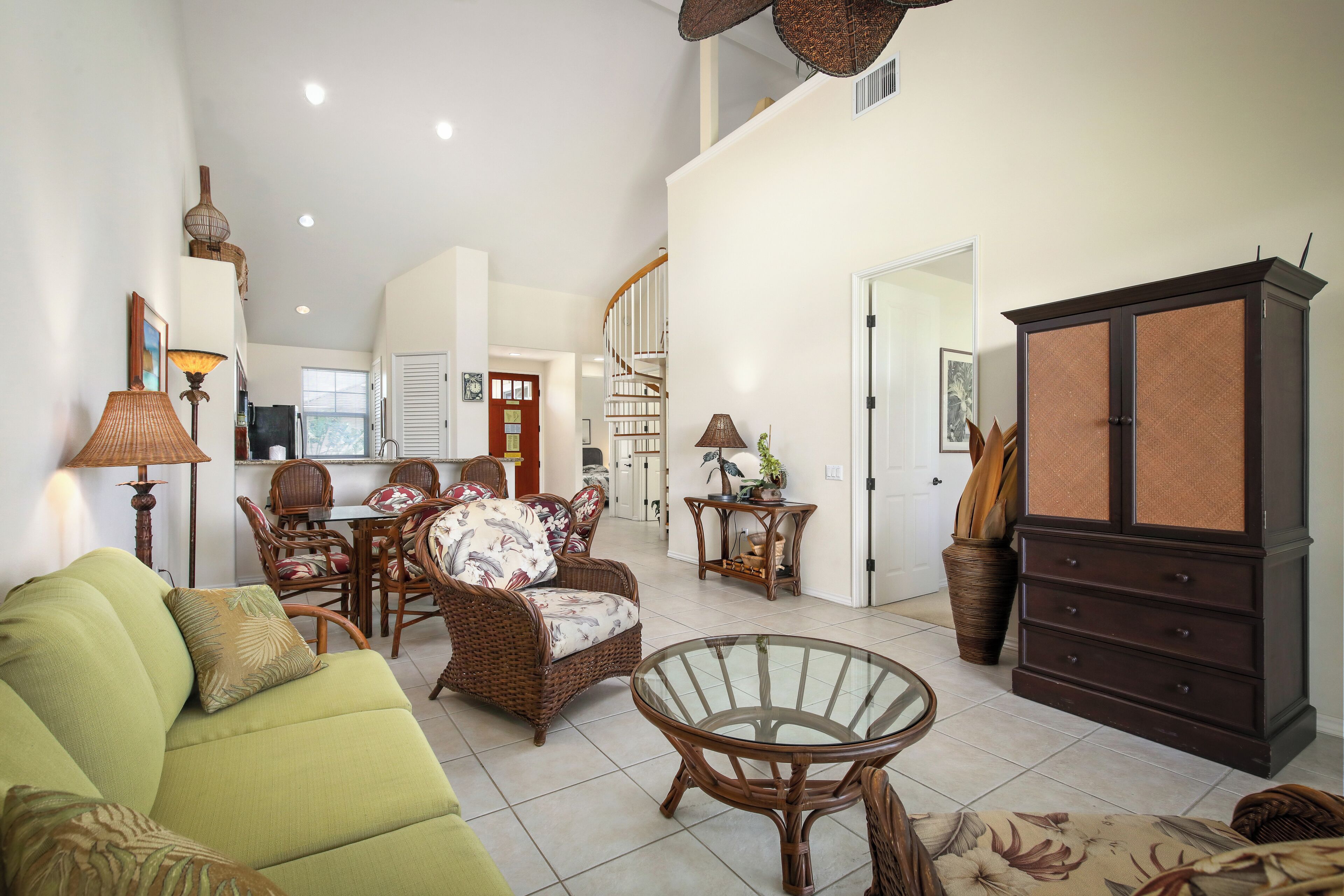 A spacious living room with a green sofa, wicker furniture, a glass coffee table, and a dark wooden cabinet.