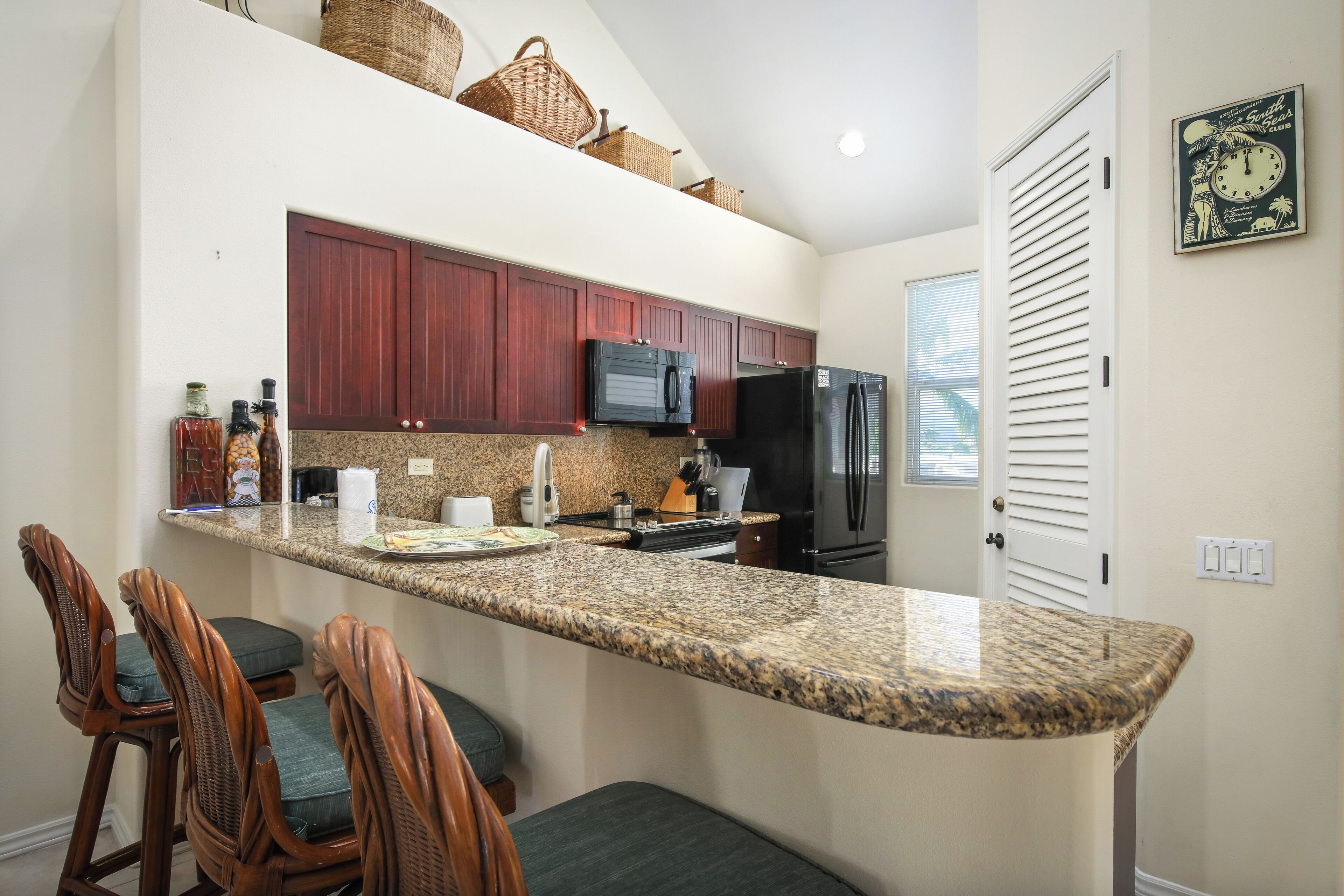 A kitchen with granite countertops, wooden chairs, a microwave, refrigerator, and a clock on the wall.