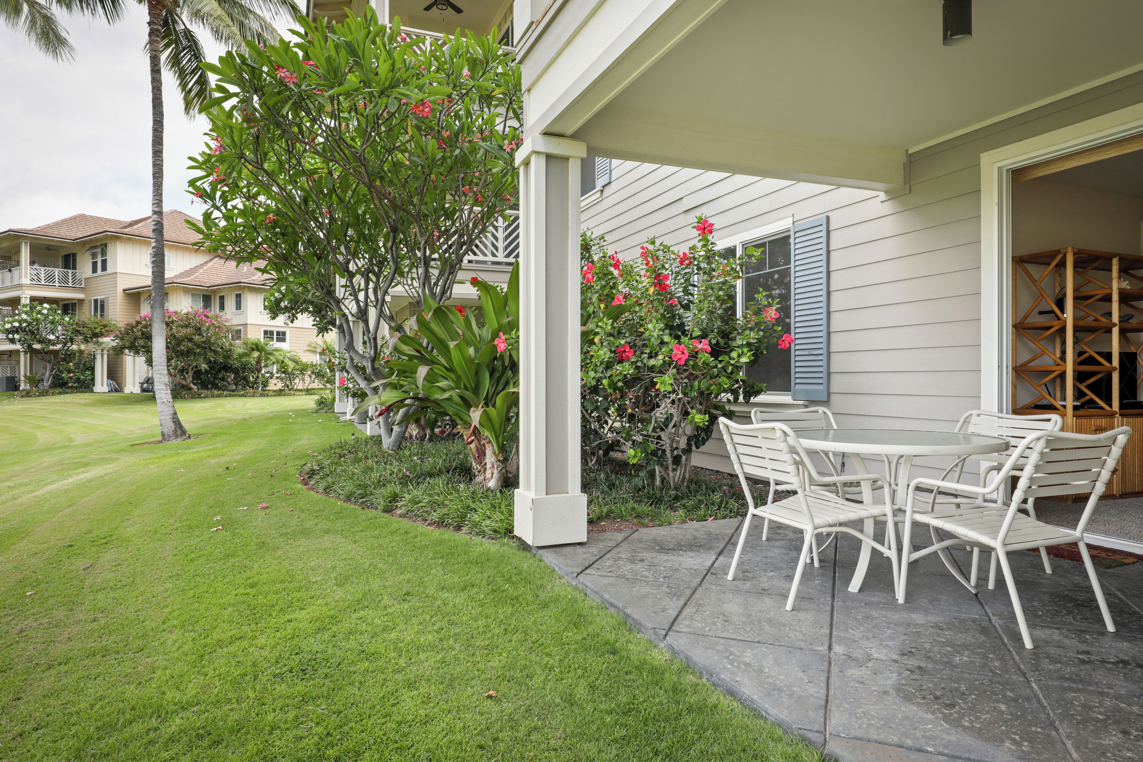A covered patio with a table and chairs, surrounded by lush greenery and flowering plants.