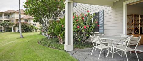 A covered patio with a table and chairs, surrounded by lush greenery and flowering plants.