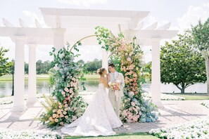 A wedding ceremony under a white pergola with floral decorations, a lake in the background, and a paved walkway.