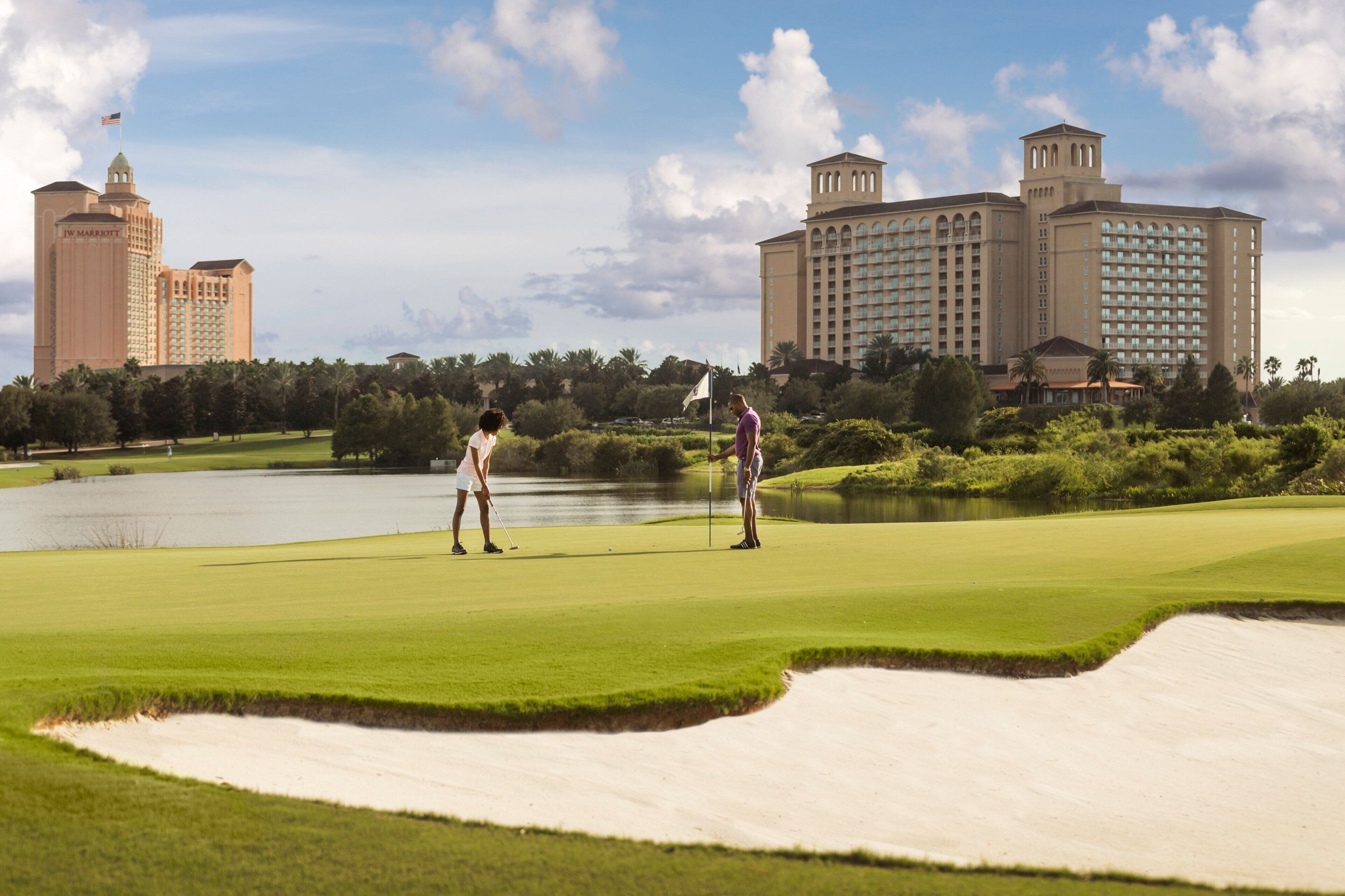Campo de golf con búnkeres de arena, un estanque y dos edificios al fondo.