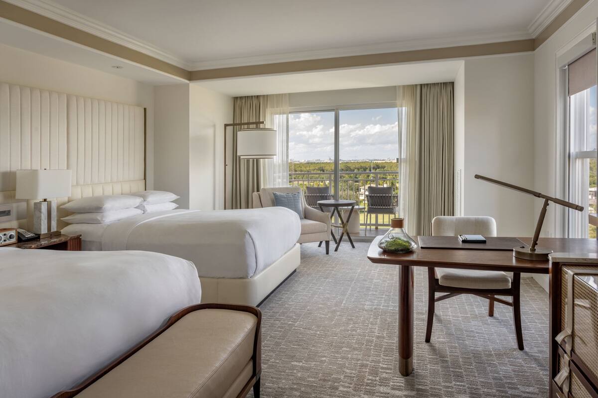 Quarto de hotel com duas camas, uma mesa, uma cadeira, um abajur e vista para o exterior.