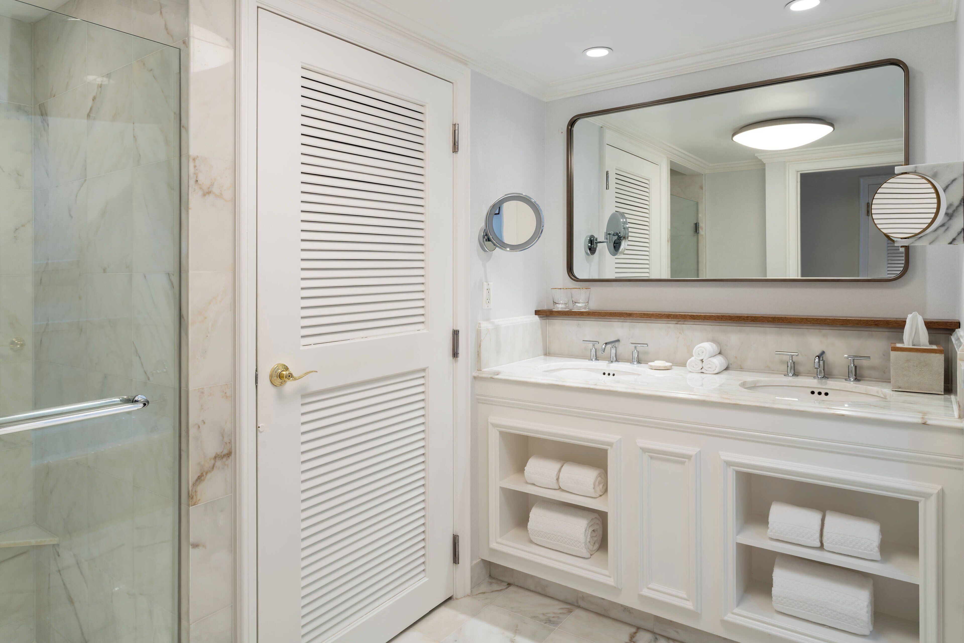 Un baño con cabina de ducha de vidrio, tocador con un espejo grande y estantes empotrados para toallas.