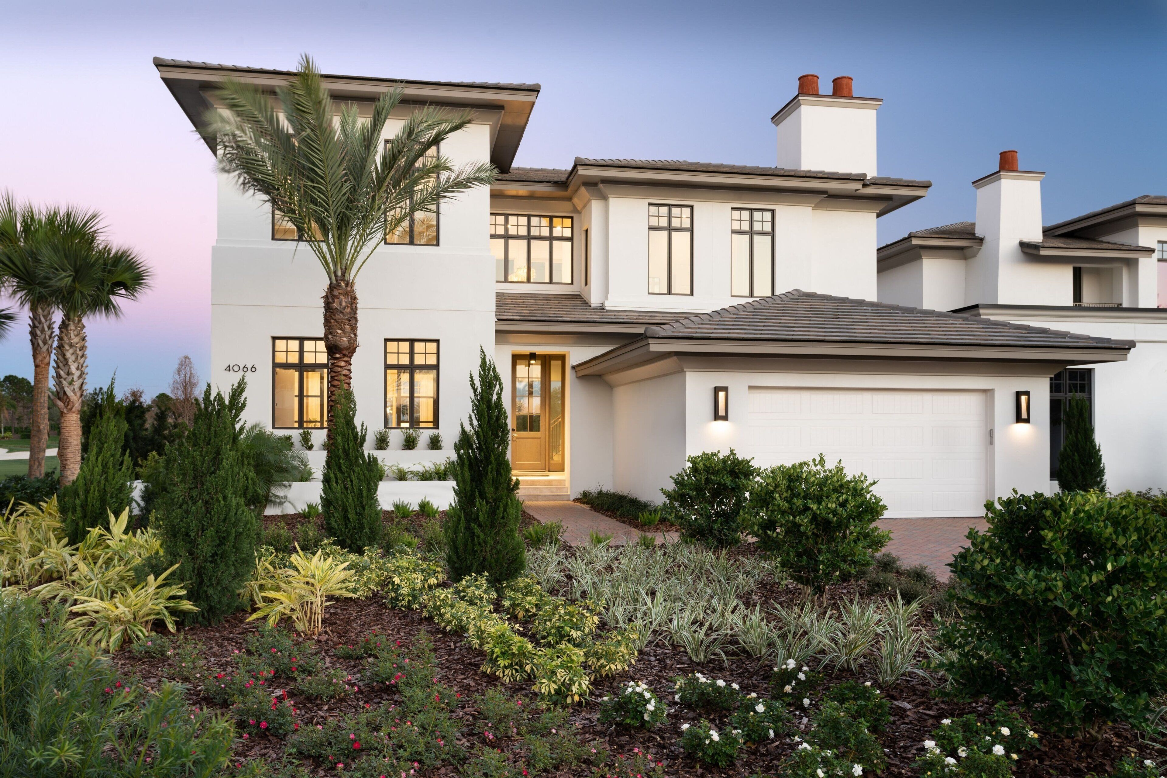 Una moderna casa a due piani con facciata bianca, un garage e un giardino curato con diverse piante e una palma.