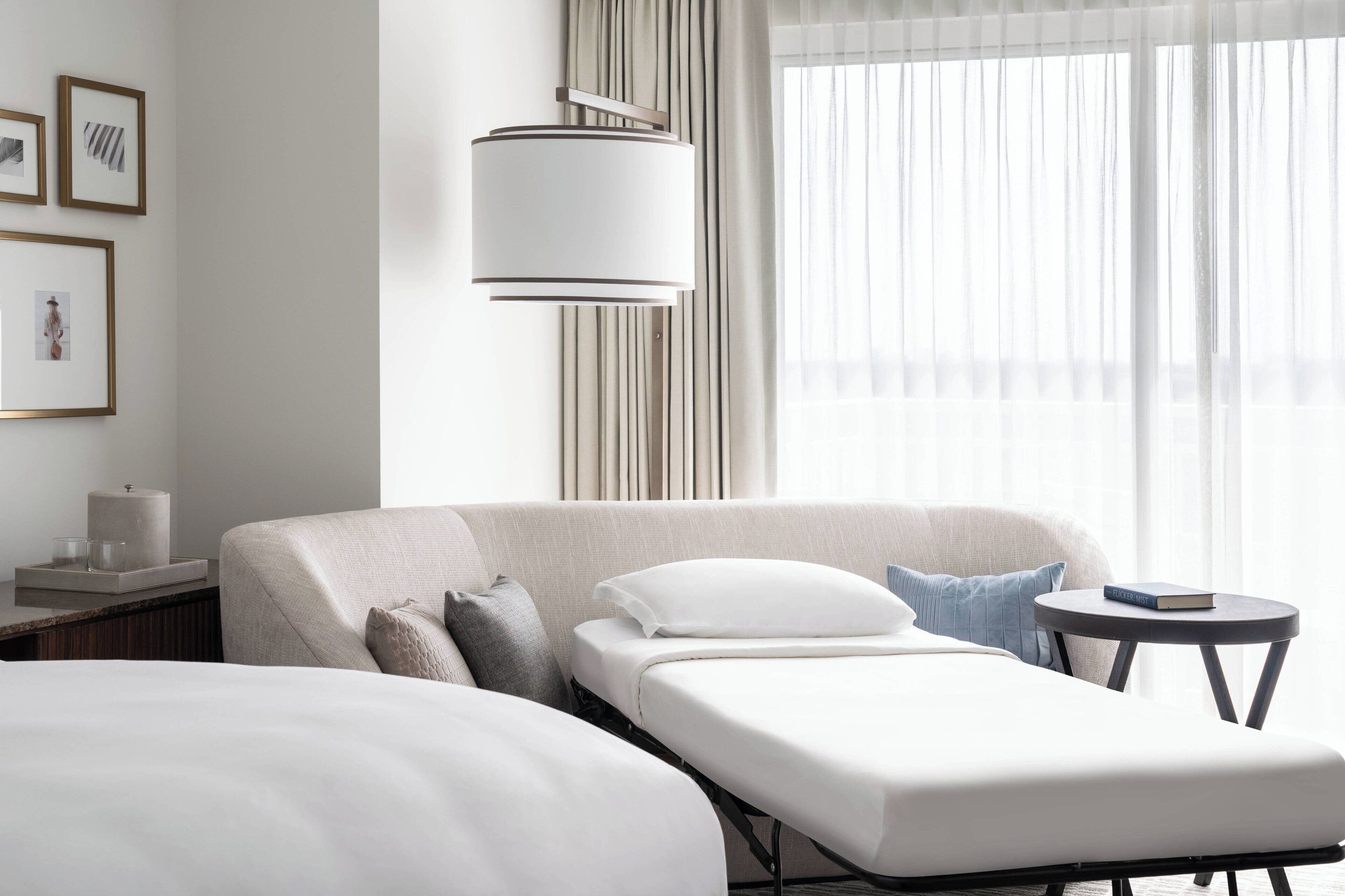 Een moderne hotelkamer met een groot bed, een nachtkastje met lamp, een klein rond tafeltje en een raam met dunne gordijnen.