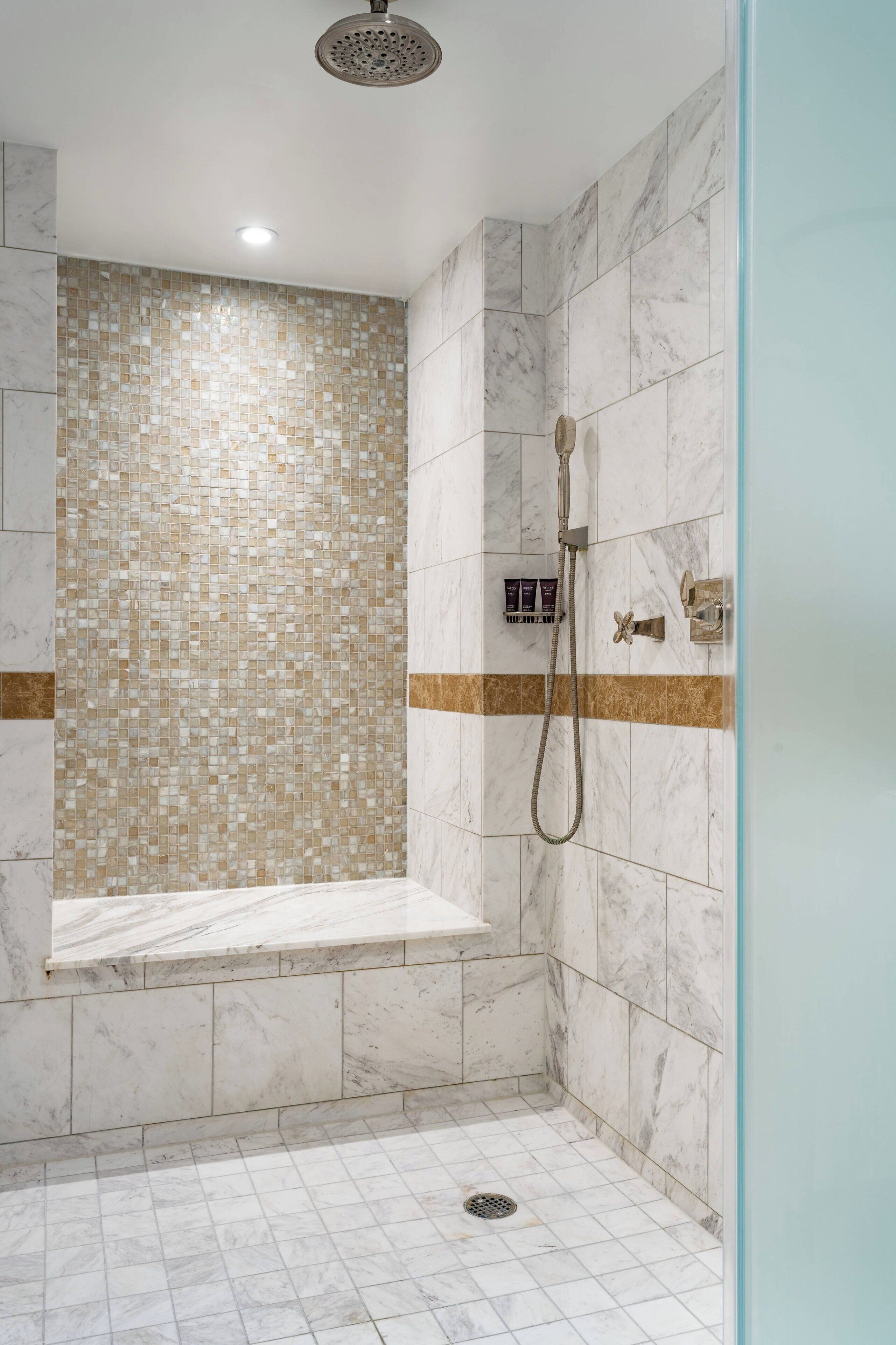 Eine Dusche mit Fliesen, einem Mosaik an der Wand, einem Handbrausekopf und einer kleinen Ablage für Pflegeprodukte.