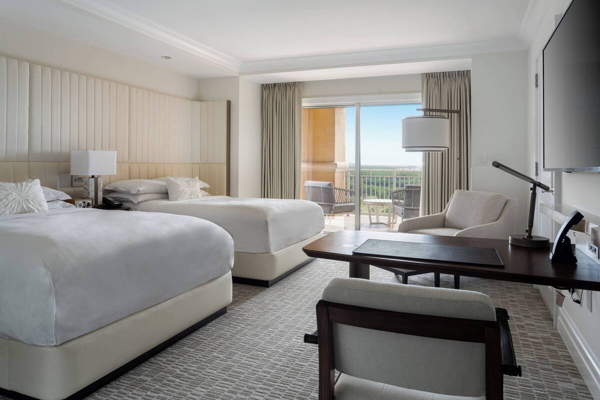 Quarto de hotel com duas camas, uma escrivaninha, uma cadeira, televisão e varanda com vista.
