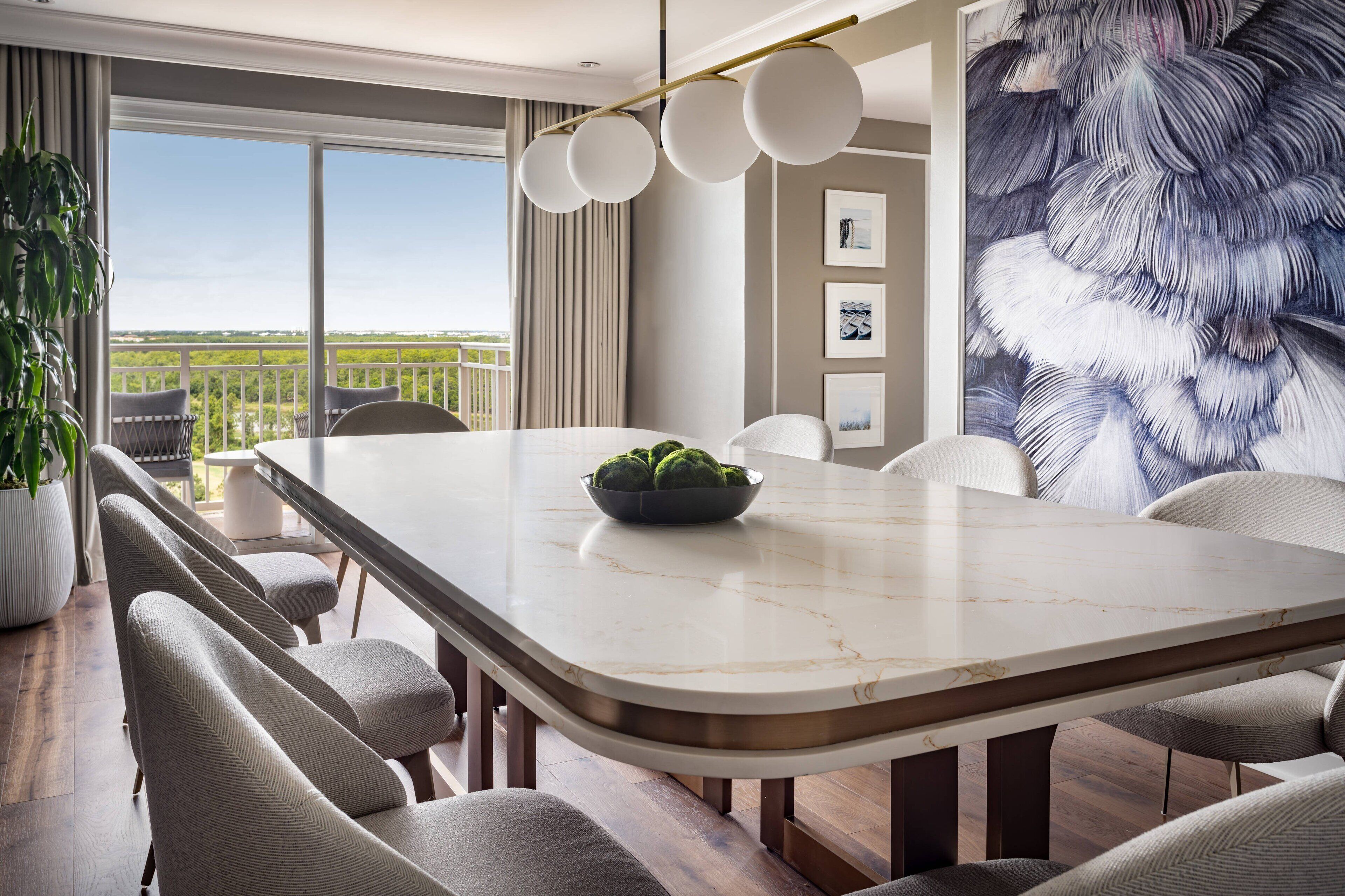 A modern dining area with a marble table, grey upholstered chairs, a potted plant, and a large window with a view.