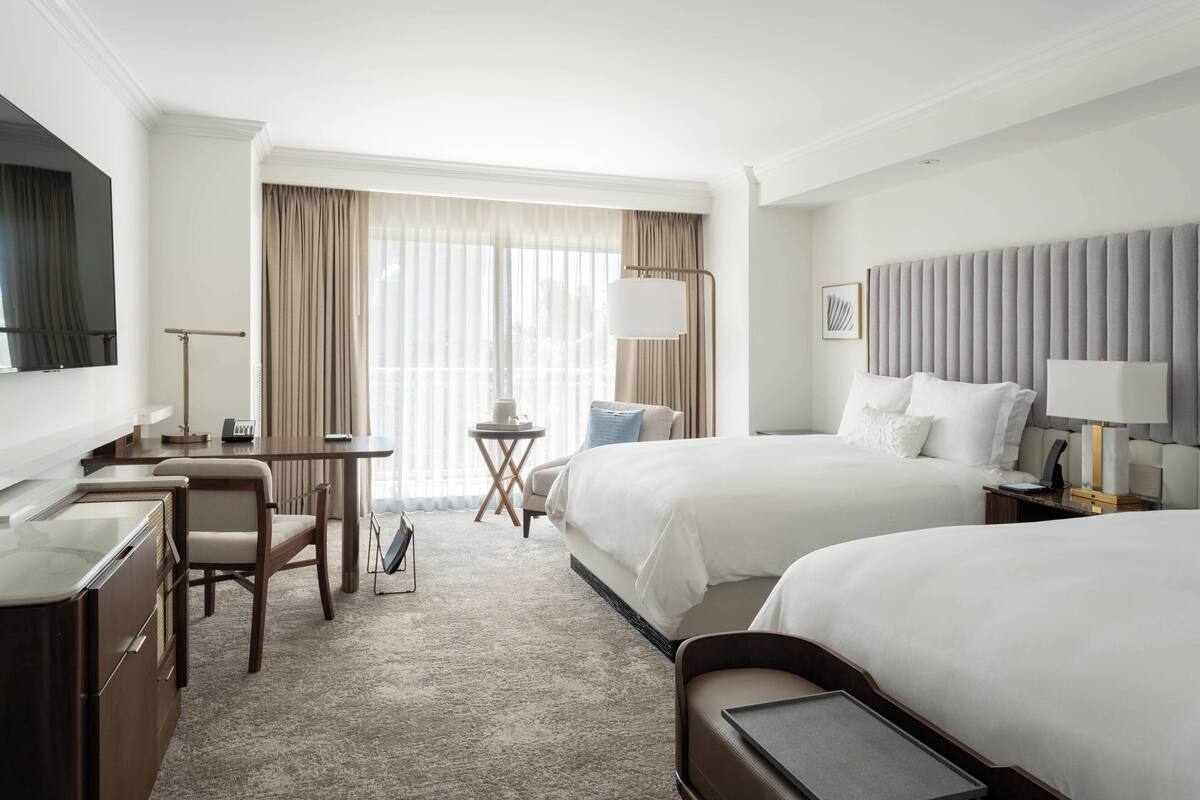 Quarto de hotel com duas camas, uma escrivaninha, uma cadeira, uma televisão e uma janela com cortinas.