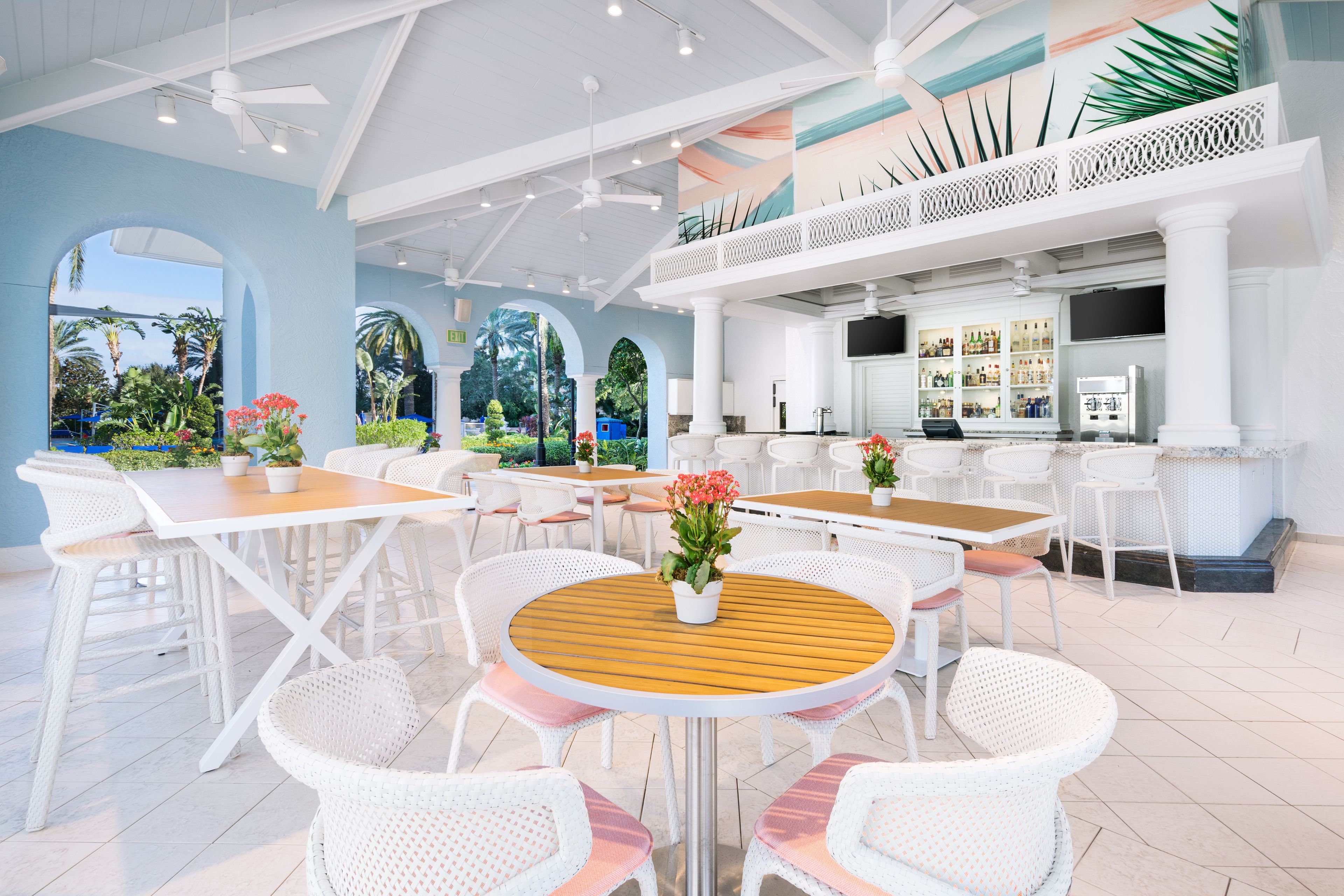 A spacious outdoor dining area with white wicker furniture and a central wooden table, surrounded by a bar area with a variety of beverages.