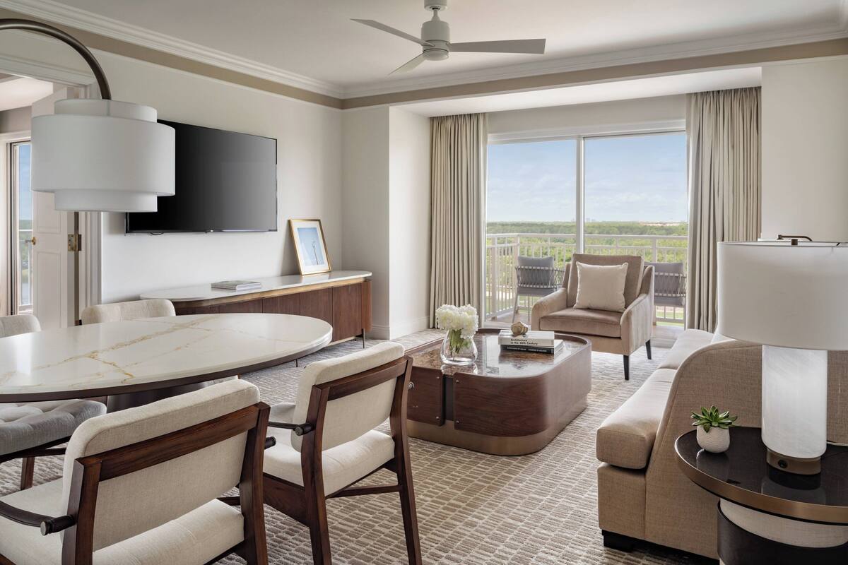 Quarto de hotel com área de refeições, sofá, mesa de centro e TV fixada na parede.