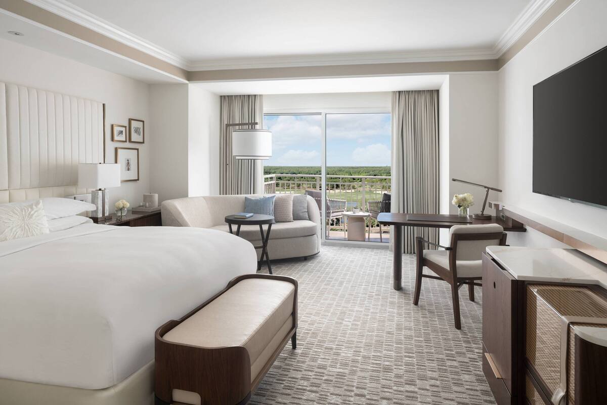 Quarto de hotel com uma cama grande, um sofá, uma escrivaninha e uma televisão. Possui uma varanda com vista para a paisagem.